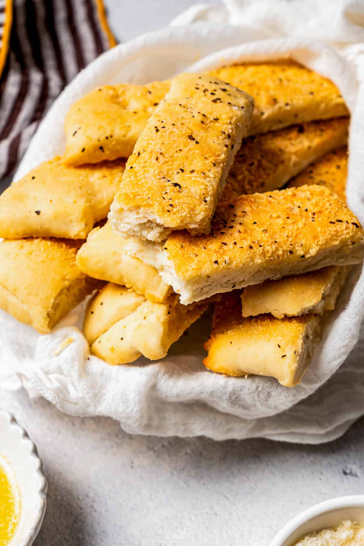 Garlic breadsticks wrapped with a white cloth, next to a small bowl of garlic dipping sauce.