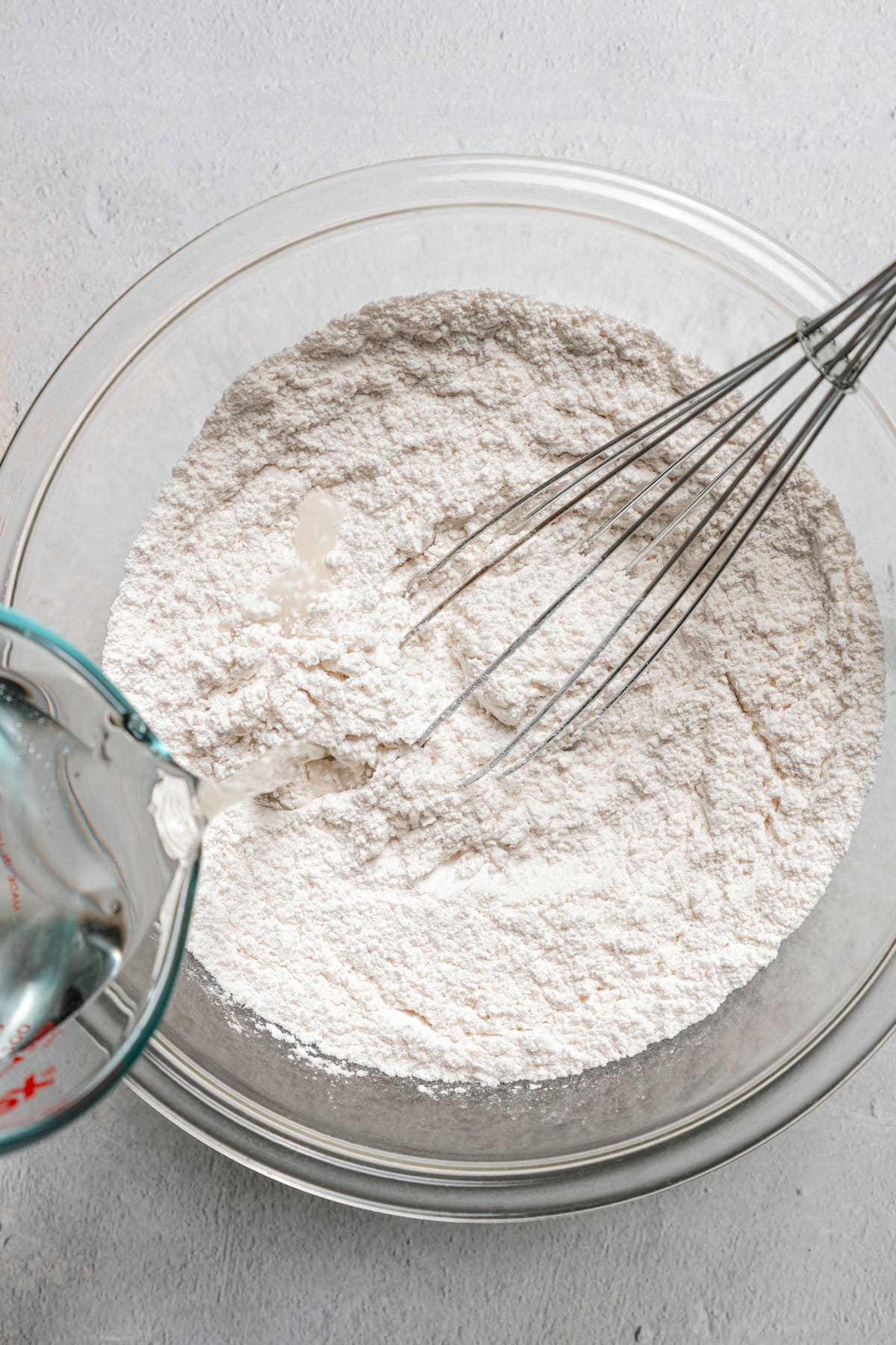 A whisk resting in a bowl of dry dough ingredients while water is being poured over top.