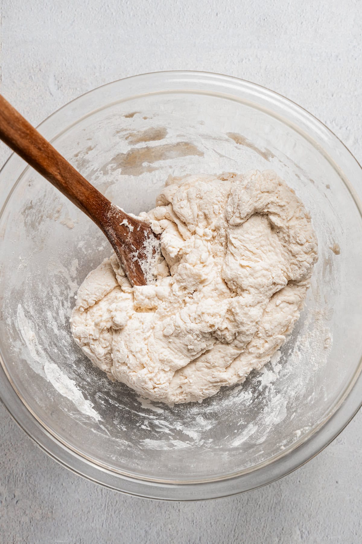 Pizza dough coming together in a glass bowl with a wooden spoon.