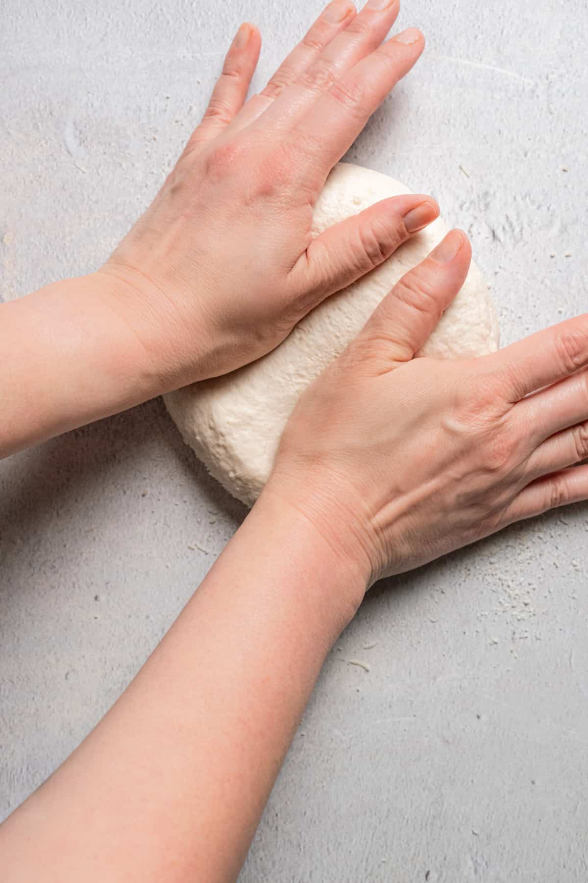 Two hands kneading a ball of yeast-free pizza dough on a clean surface.