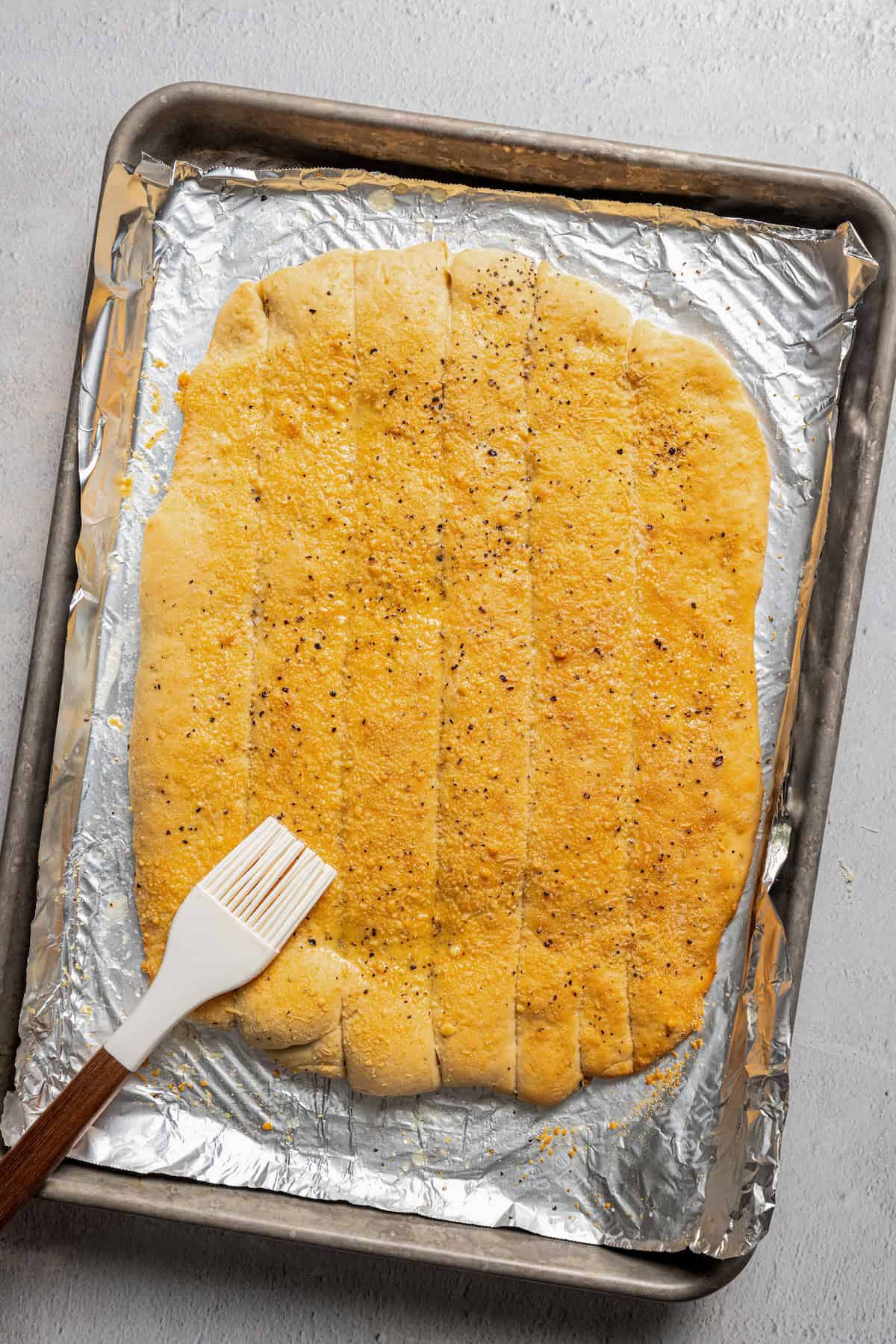 Garlic breadsticks sprinkled with parmesan cheese being brushed with garlic butter on a foil-lined baking tray.