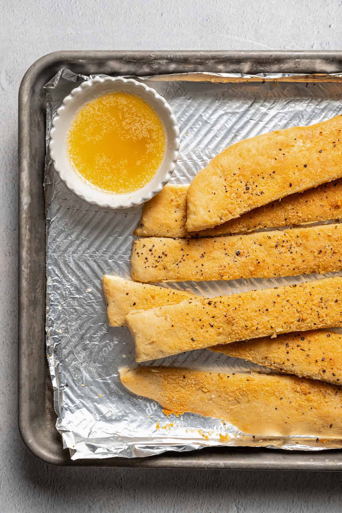 Garlic parmesan breadsticks on a foil-lined baking tray next to a bowl of garlic dipping sauce.