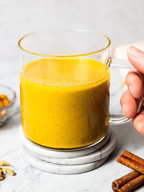 A hand holding a glass mug filled with turmeric latte near a bowl of turmeric and cinnamon sticks.