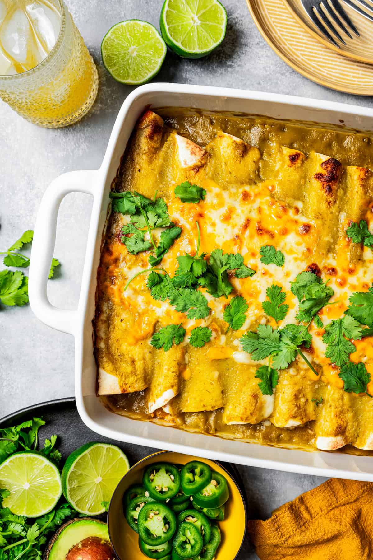 Overhead view of green chili chicken enchiladas in a baking dish garnished with fresh clantro leaves, next to bowls of lime halves and sliced jalapeños.