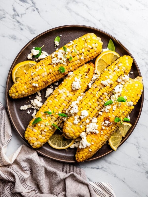 Overhead view of four ears of corn on a plate topped with goat cheese and garnished with lemon wedges.