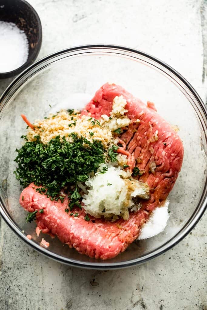 a glass mixing bowl with ingredients for turkey burgers; ground turkey, chopped herbs, seasonings, onions, mustard, and worcestershire sauce