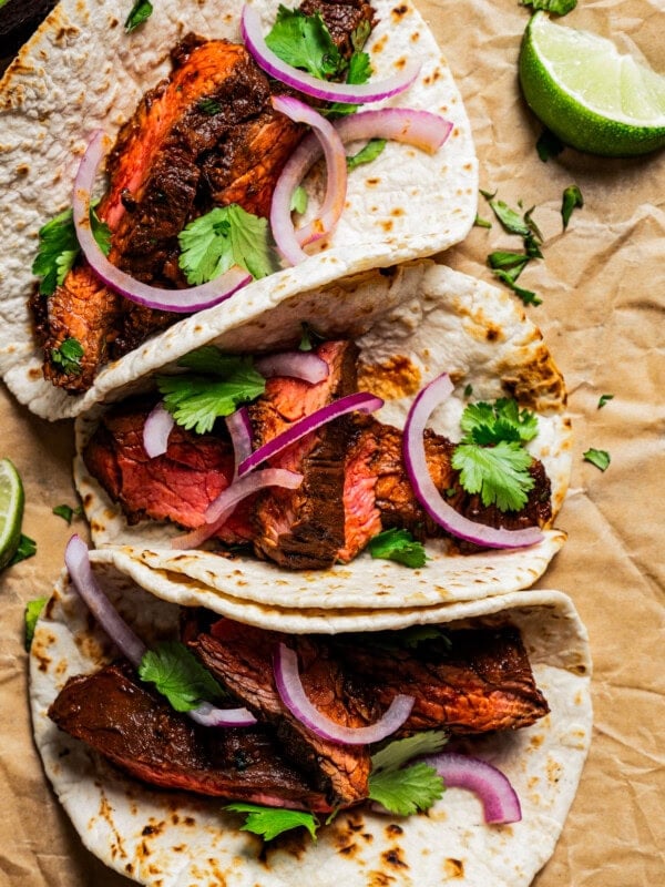 Overhead view of three garlic guajillo steak tacos garnished with sliced onions and cilantro, next to scattered lime wedges.