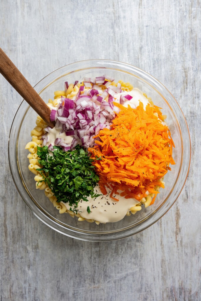 Adding carrots, dressing, red onion, and parsley to a bowl of cooked macaroni.