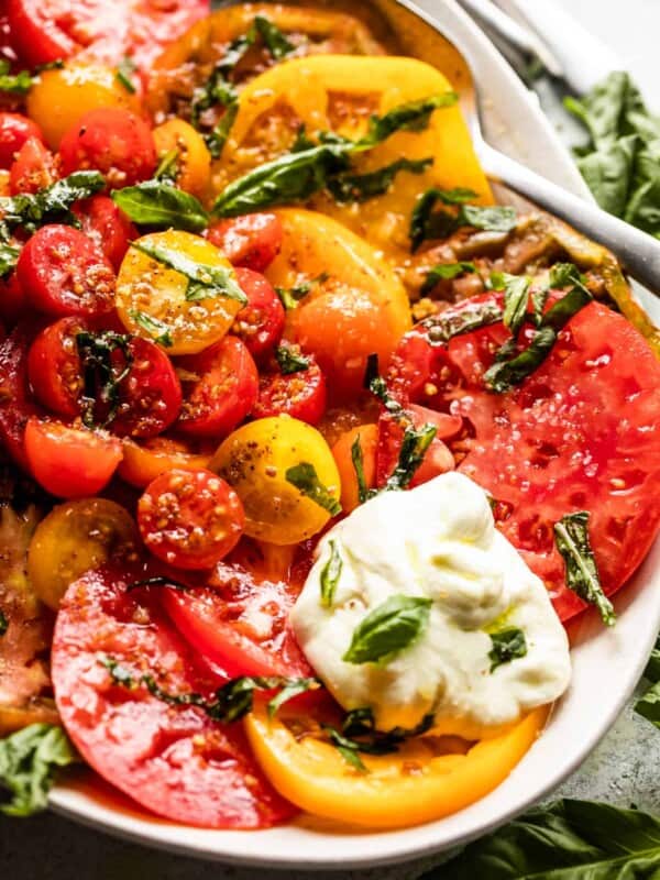 Up close shot of Tomato Burrata Salad: slices of heirloom tomatoes arranged on an oval platter, with halved cherry tomatoes placed in the center, burrata cheese at the top of the plate and the bottom, and a garnish of basil over the entire salad.