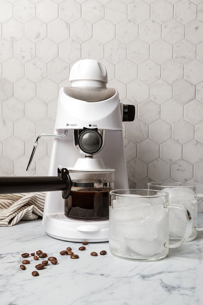 Two glasses filled with iced next to an espresso machine brewing espresso on a white marble countertop.