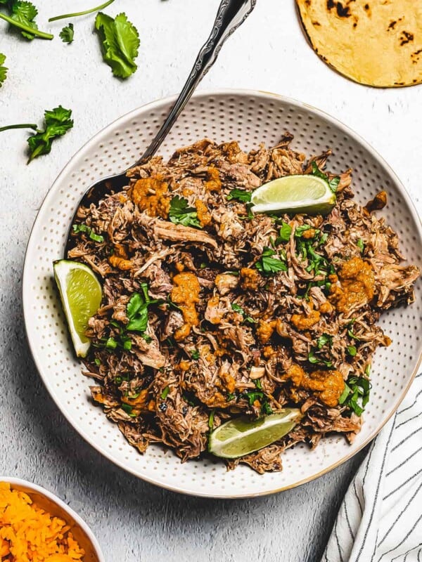 overhead shot of shredded pork pernil in a white serving dish, and garnished with lime slices plus parsley. Corn tortillas are placed to the right, near the serving dish, and a bowl of rice is placed on the opposite side.