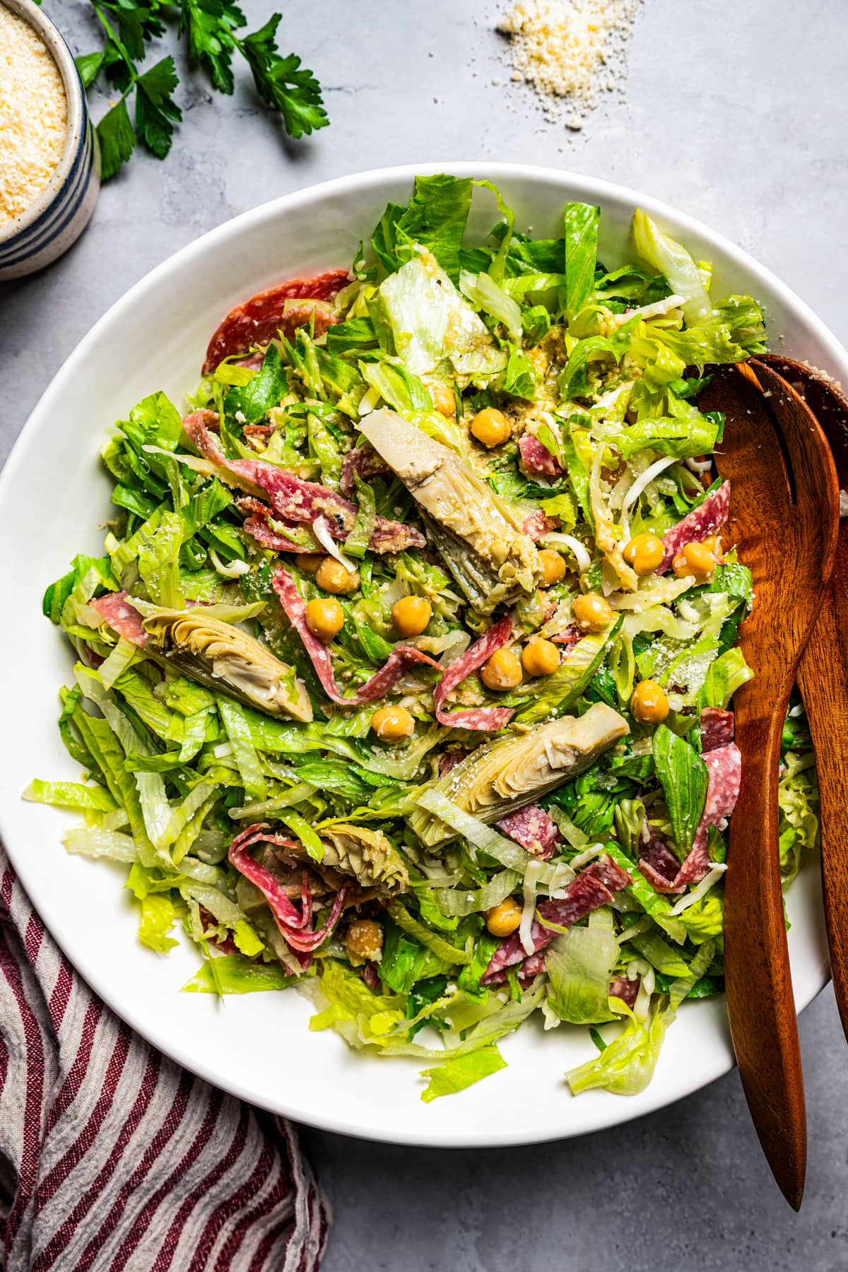 Overhead view of La Scala chopped salad in a large bowl with sliced artichokes, salami, and chickpeas, next to two wooden serving spoons.