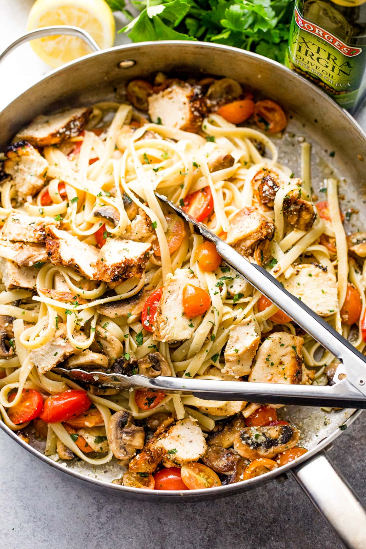 Pair of tongs tossing chicken and pasta with cherry tomatoes and mushrooms in a skillet.