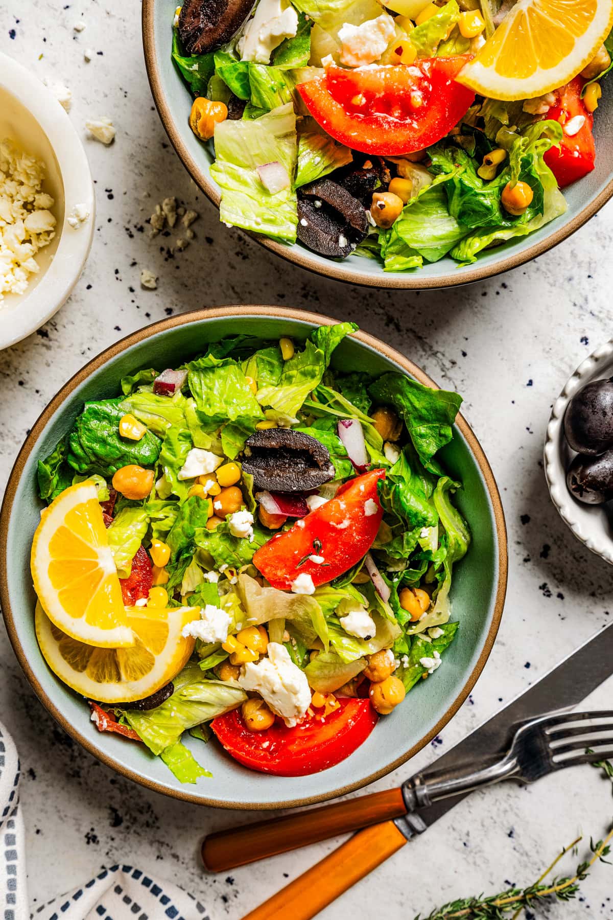 Two bowls filled with romaine lettuce, tomatoes, corn, chickpeas, and slices of lemon.