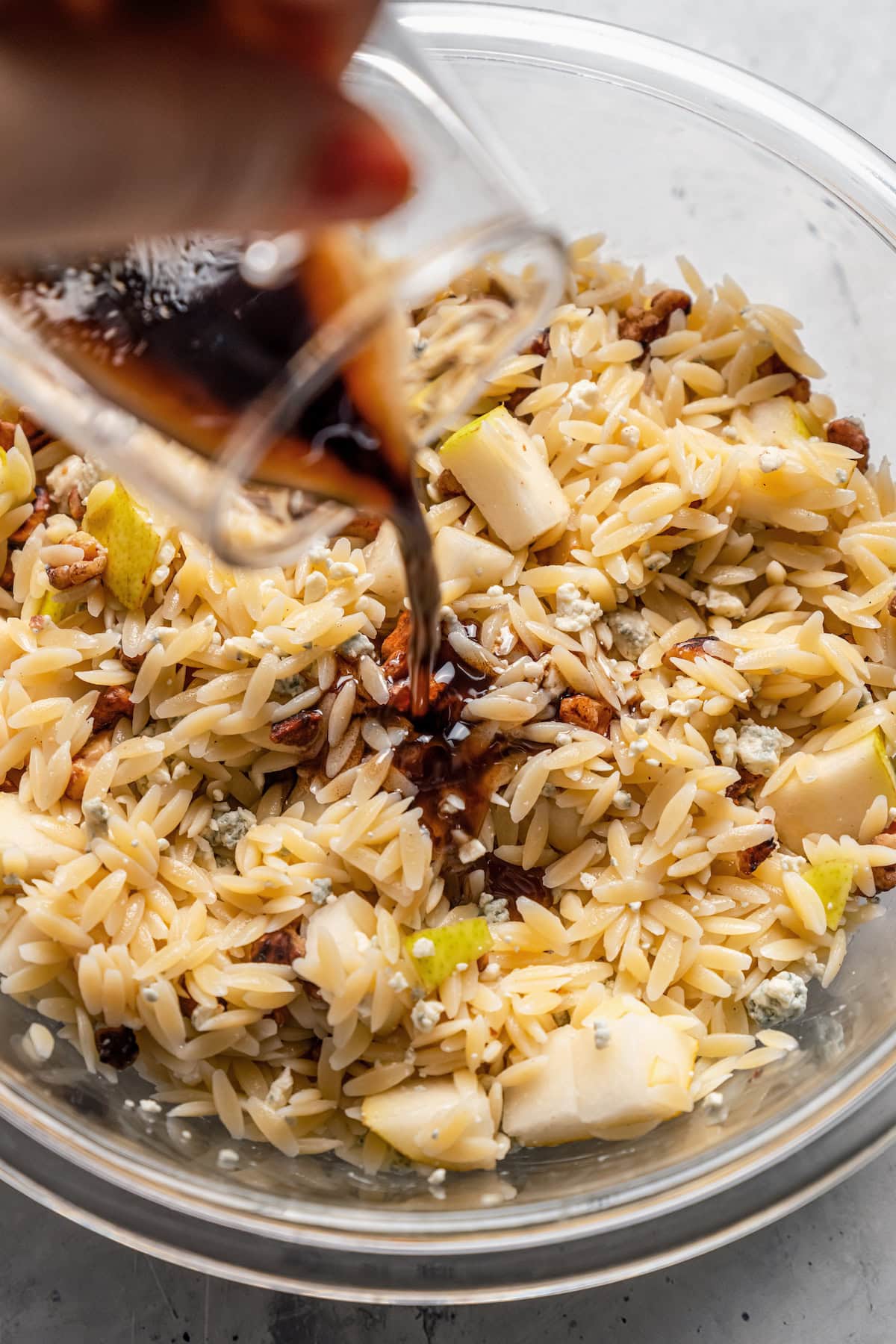 Balsamic dressing being poured over orzo salad ingredients in a glass bowl.