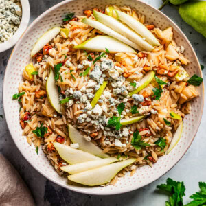 Overhead view of orzo salad with pears, walnuts and gorgonzola cheese in a bowl garnished with pear slices, with a wooden spoon, surrounded by bowls of ingredients.