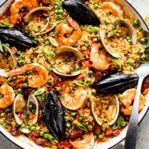 Spanish paella in a white braiser: arborio rice topped with clams, shells, mussels, shrimp, sliced sausages, and peas.