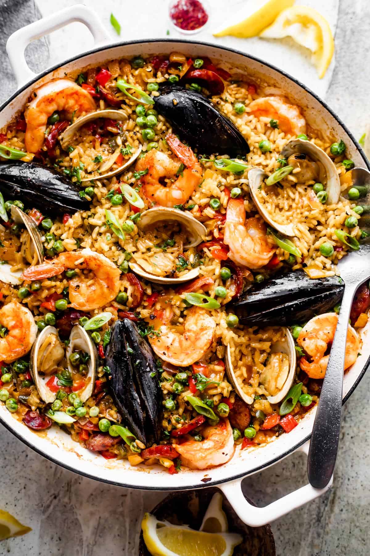 Spanish paella in a white braiser. Photo includes arborio rice topped with clams, shells, mussels, shrimp, sliced sausages, and peas.