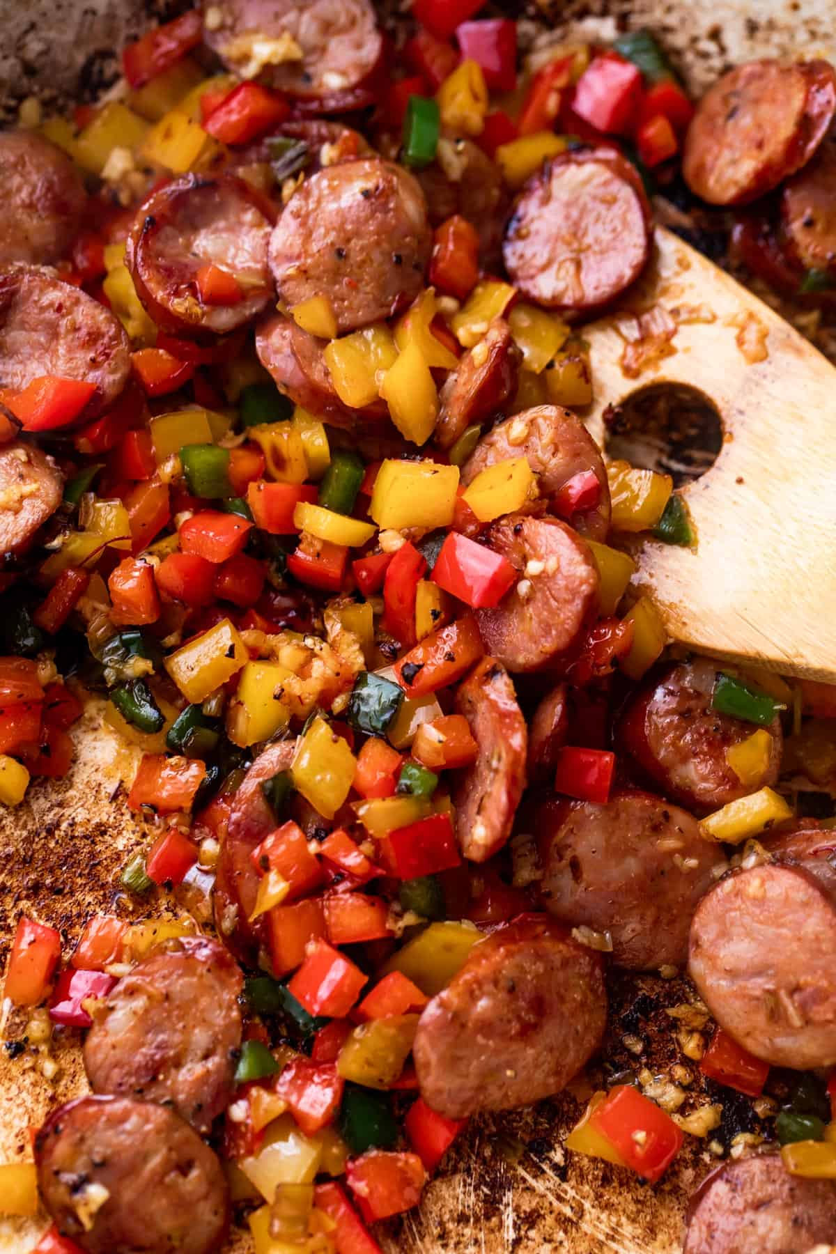 Wooden spoon mixing through sliced pork sausages and diced bell peppers.