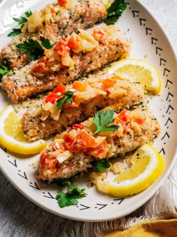 Panko-crusted salmon filets served on a platter next to lemon wedges and topped with Tuscan tomato sauce.