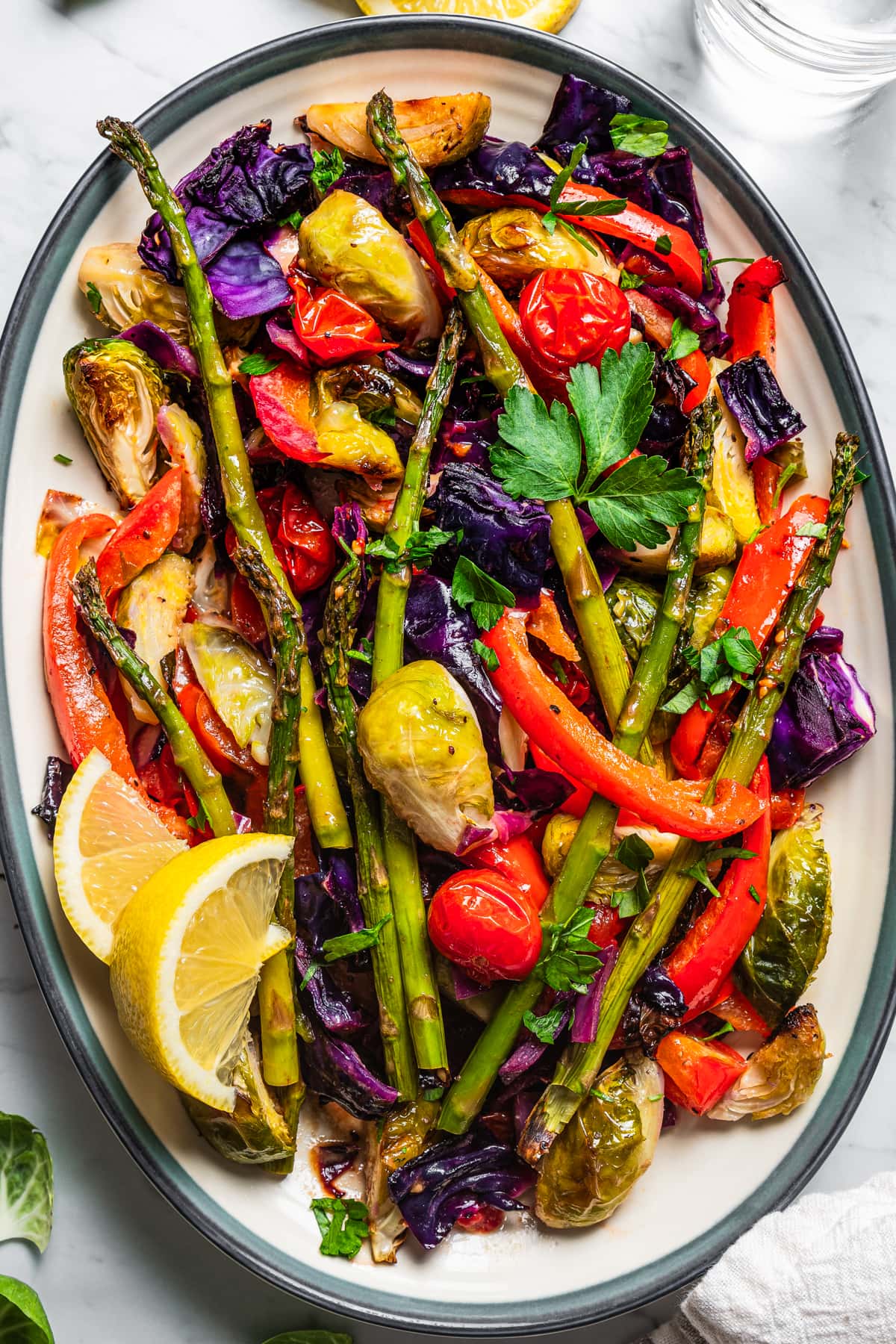 Roastd asparagus, bell peppers, tomatoes, and brussel sprouts served on a platter with lemon wedges.