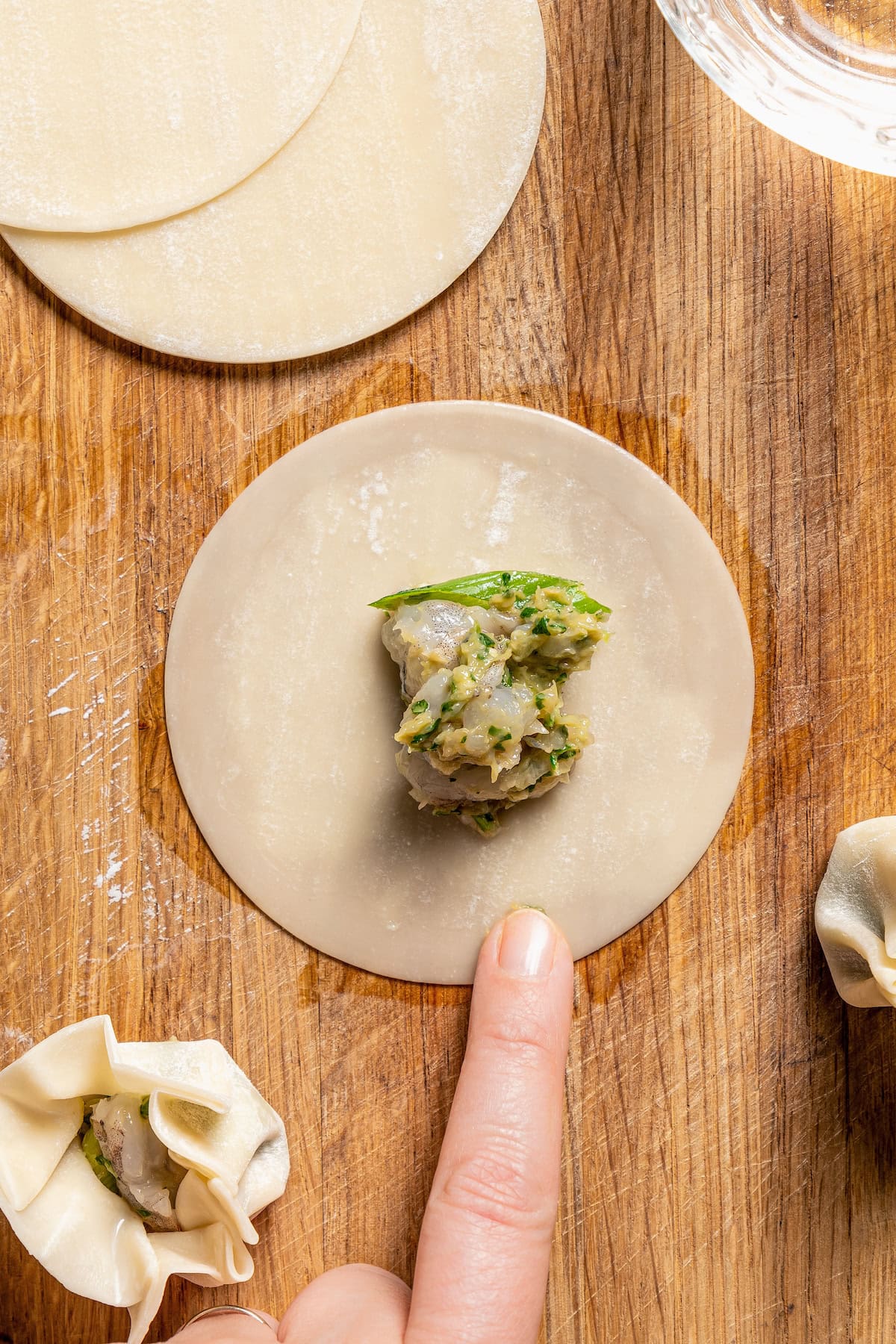 A spoonful of shrimp filling in the center of a round dumpling skin, while a finger rubs the edge with water.