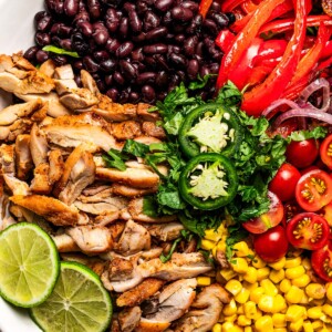 Close-up shot of a bowl with sliced chicken, sliced red bell peppers, black beans, tomatoes, and corn.