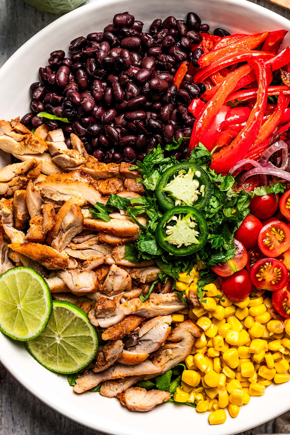 Close-up shot of a bowl with sliced chicken, sliced red bell peppers, black beans, tomatoes, and corn.