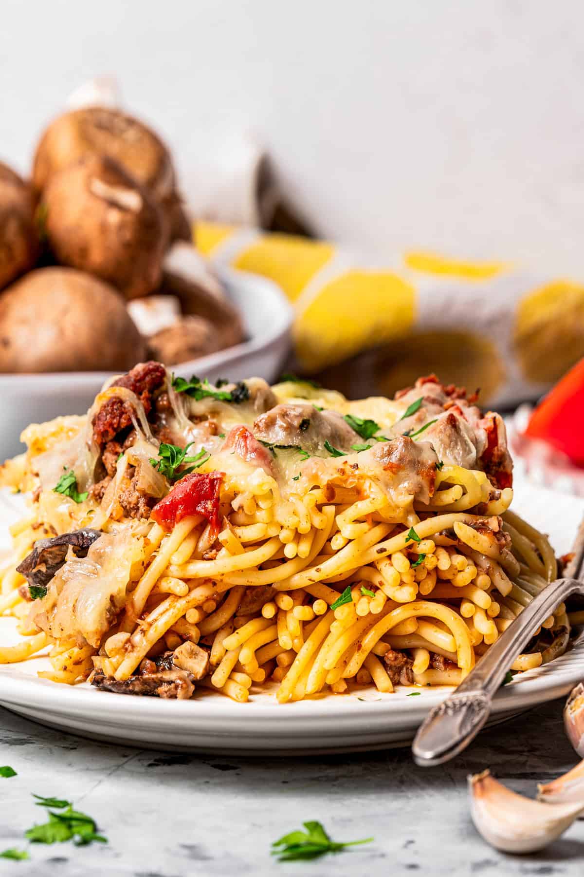 A serving of spaghetti casserole on a plate next to a fork, with a bowl of mushrooms next to a quartered tomato in the background.