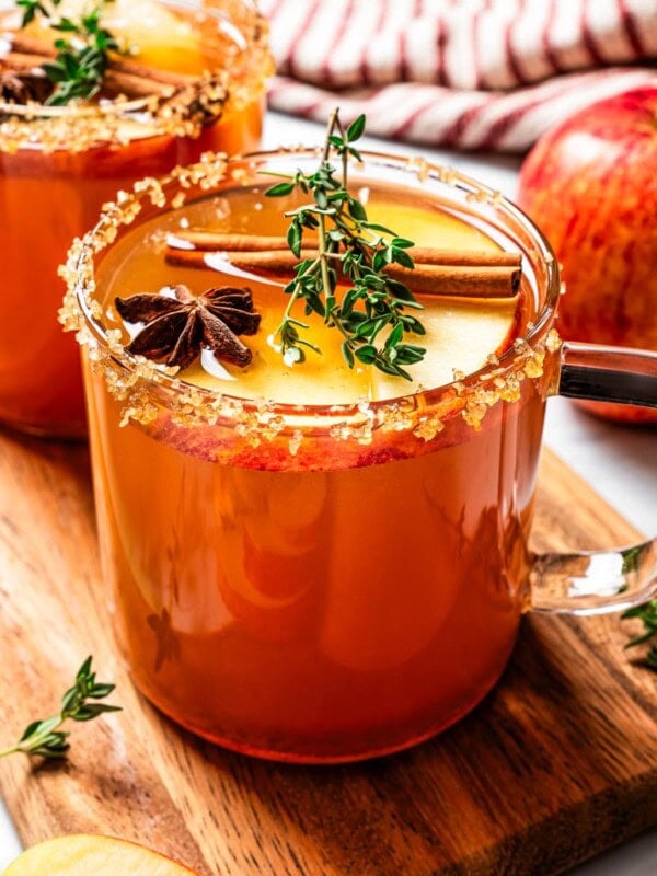 A glass cup filled with spiked apple cider garnished with apple slices, thyme, anise, and a cinnamon stick.