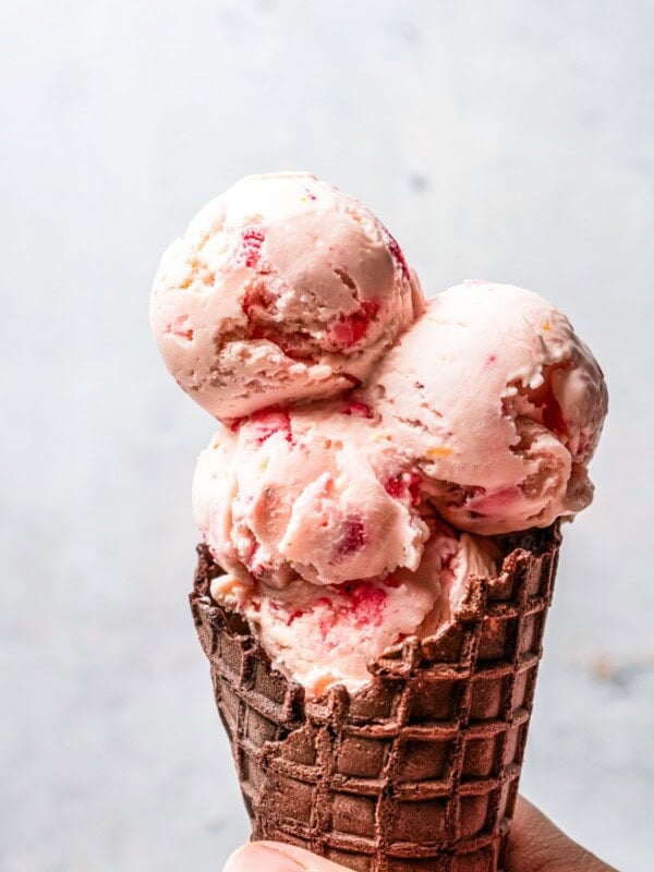 Hand holding up a waffle cone filled with three scoops of strawberry cheesecake ice cream.