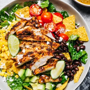 A large bowl with a salad featuring sliced chicken breast, cherry tomatoes, nacho chips, avocado, onions, black beans, and corn.