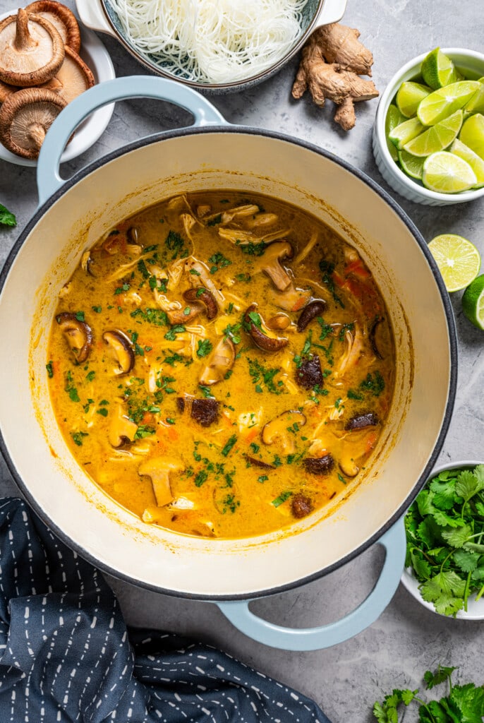 Thai chicken soup in a Dutch oven surrounded by bowls of lime slices, rice noodles, mushrooms, and herbs.