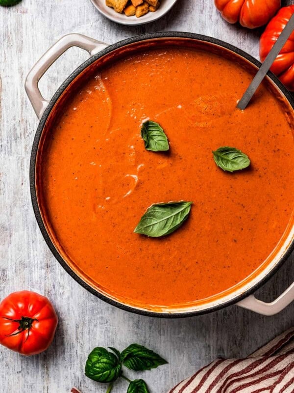 overhead shot of a soup pot with tomato bisque, topped with three basil leaves, and tomatoes and croutons placed around the pot.