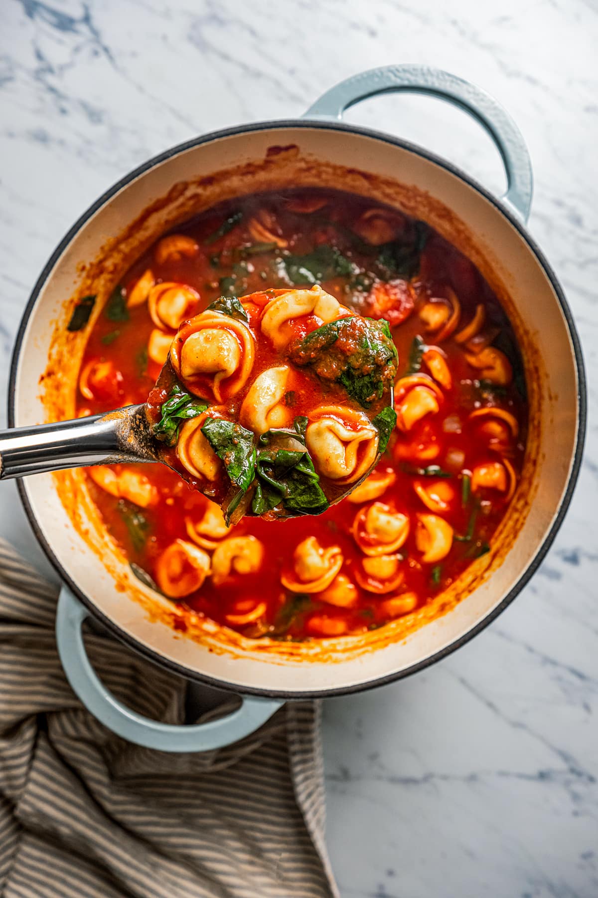 A ladle holding up spinach tortellini soup over a pot.