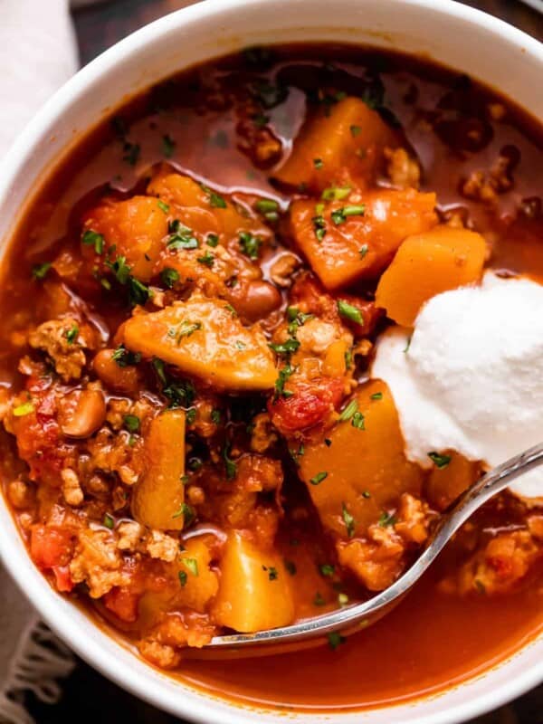 close up shot of a bowl with turkey chili, studded with cubed butternut squash, tomatoes, and a garnish of sour cream.