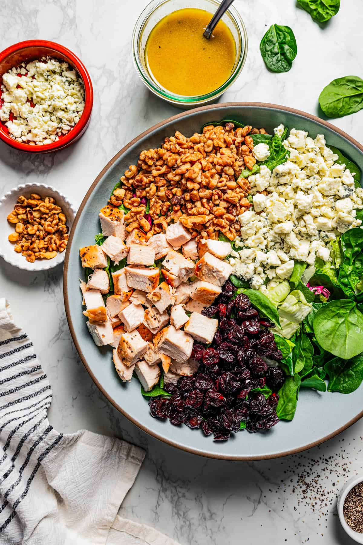 Overhead view of walnut chicken salad ingredients assembled in a large blue salad bowl, next to a jar of dressing and smaller bowls of toppings.