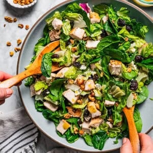 Two hands using wooden salad tongs to toss the chicken salad with the dressing.