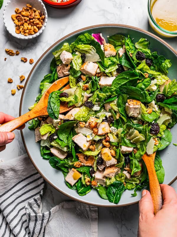 Two hands using wooden salad tongs to toss the chicken salad with the dressing.