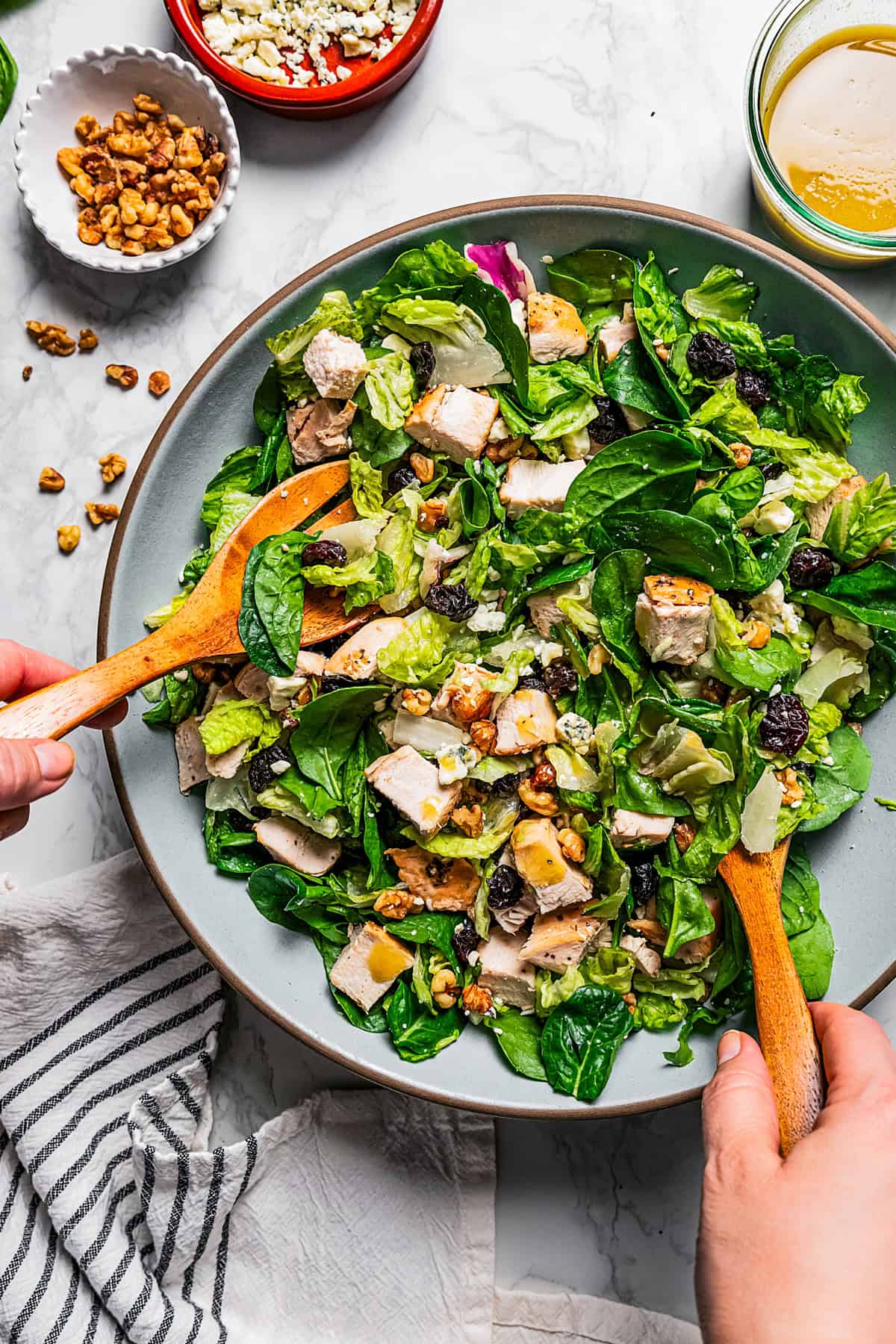 Two hands using wooden salad tongs to toss the chicken salad with the dressing.