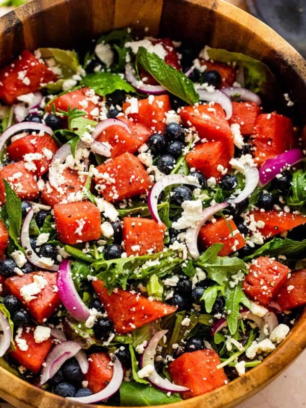 wooden salad bowl with cubed watermelon, blueberries, and sliced red onions atop a bed of torn up lettuce greens and topped with feta cheese crumbles