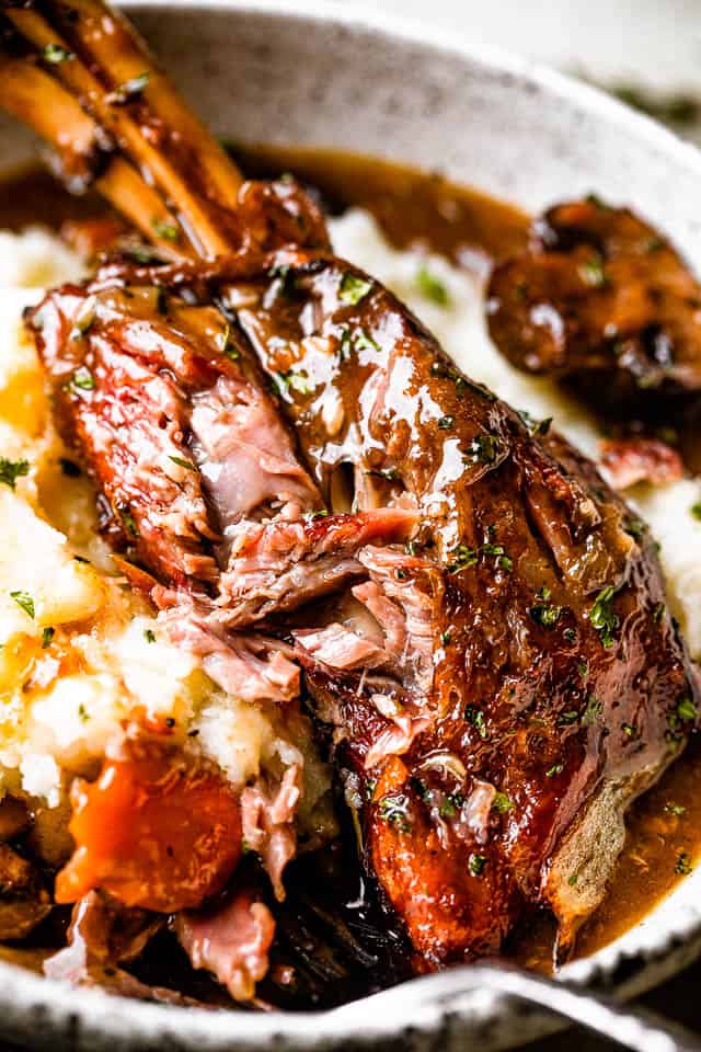 Lamb shank is served on a dinner plate with mashed potatoes, and a fork is tearing through the tender meat.