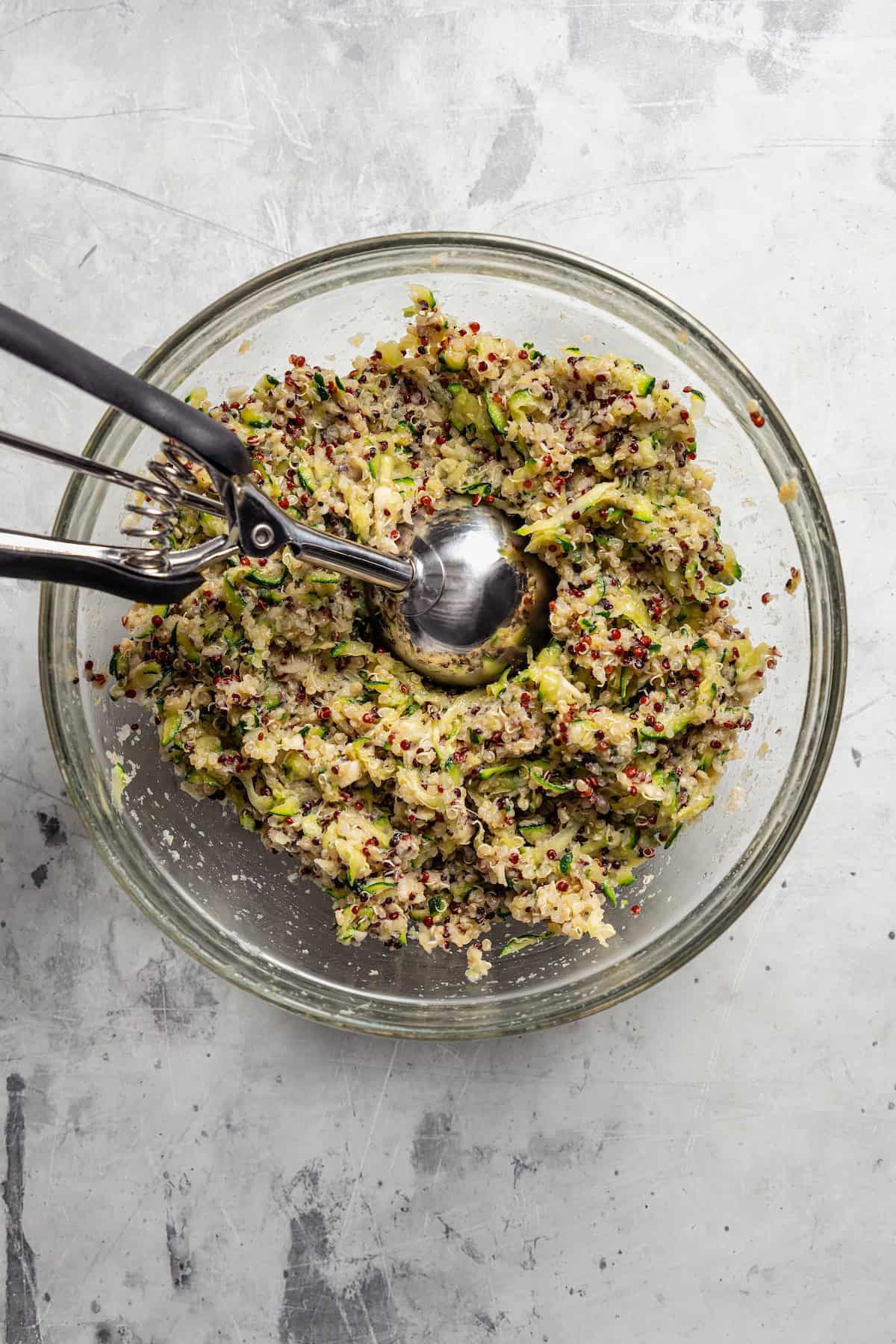 Zucchini fritter mixture in a glass bowl with a large cookie scoop.