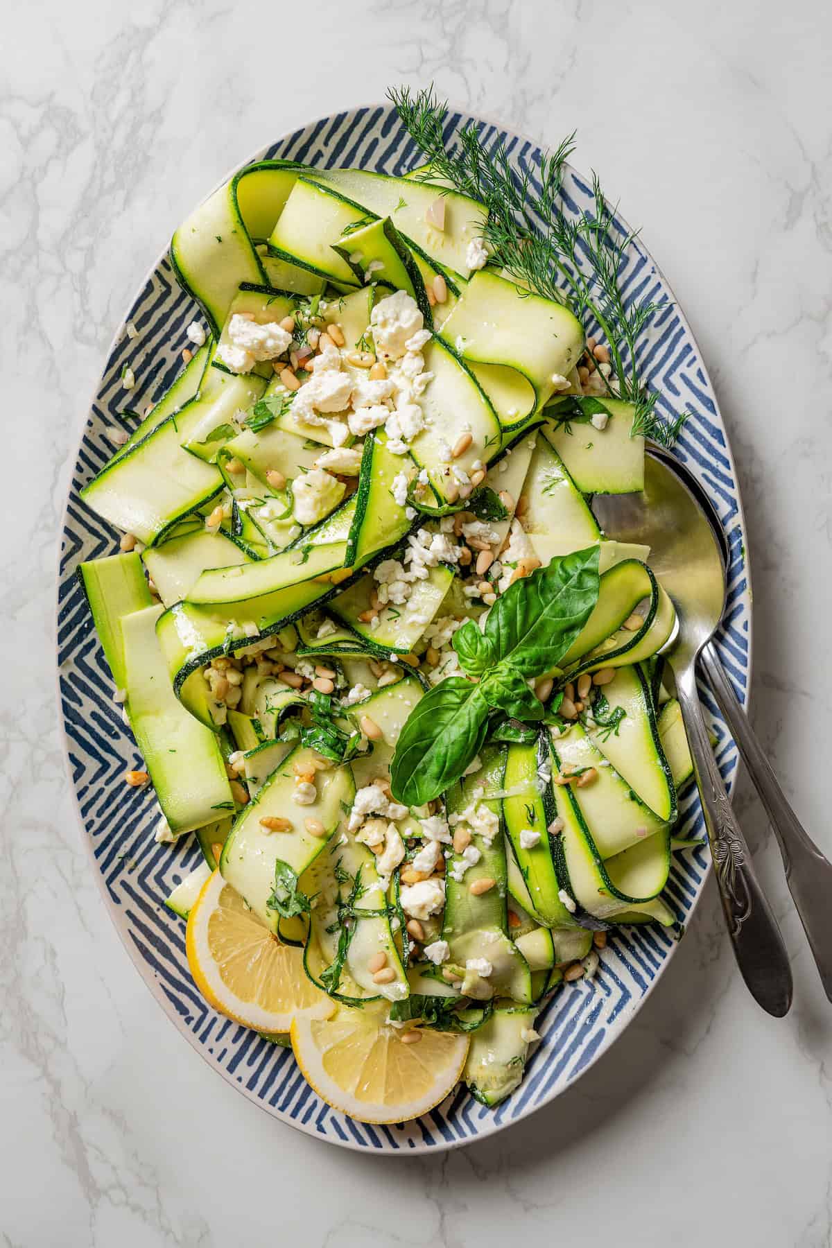 Overhead view of an assembled zucchini salad on a large platter garnished with fresh basil leaves and lemon wedges.