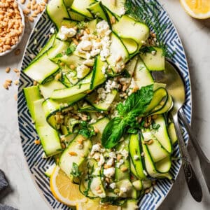 Overhead view of a salad made of zucchini ribbons and served on a large platter garnished with fresh basil leaves and lemon wedges, next to a set of salad tongs.