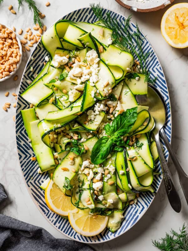 Overhead view of a salad made of zucchini ribbons and served on a large platter garnished with fresh basil leaves and lemon wedges, next to a set of salad tongs.