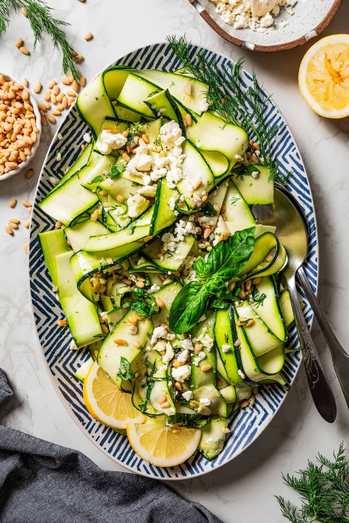 Overhead view of a salad made of zucchini ribbons and served on a large platter garnished with fresh basil leaves and lemon wedges, next to a set of salad tongs.